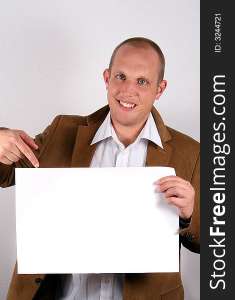 A young businessman is smiling and holds a sign and points at it! White sign to write on!. A young businessman is smiling and holds a sign and points at it! White sign to write on!