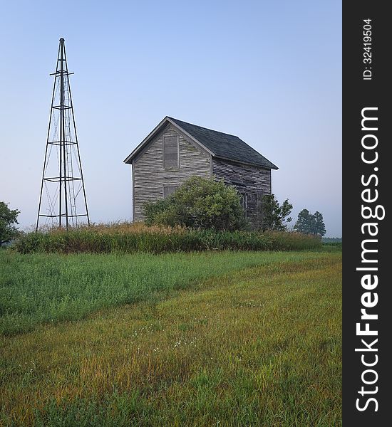 An old farm house and a windmill tower are still standing in a hay field.