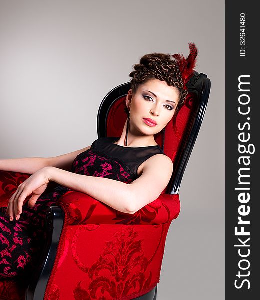 Beautiful adult woman with fashion hairstyle and red armchair poses at studio