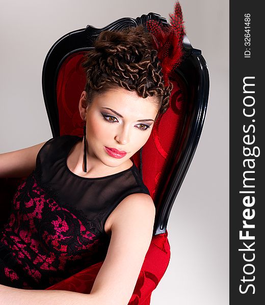 Beautiful adult woman with fashion hairstyle and red armchair poses at studio