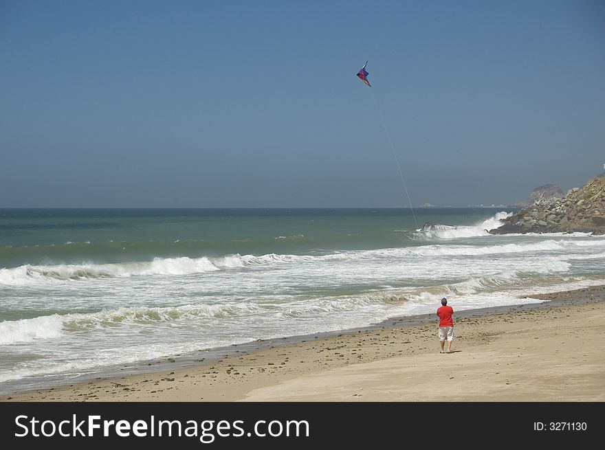 A man in a read shirt and white shorts flying a kite at the beach on the California coastline with the ocean and the blue sky in the background