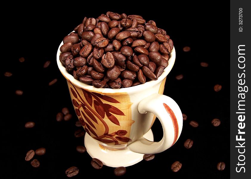 A decorative mug full of rich, dark coffee beans with scattered beans on the table around the it. Black background. A decorative mug full of rich, dark coffee beans with scattered beans on the table around the it. Black background.