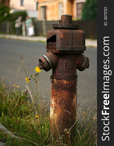 An old rusty hydrant stand pipe with a few houses and two chatting ladies on the street in the background. An old rusty hydrant stand pipe with a few houses and two chatting ladies on the street in the background.