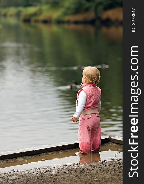 Small girl toddler dressed in pink standing in a mud puddle watching the ducks swim at the park. Small girl toddler dressed in pink standing in a mud puddle watching the ducks swim at the park