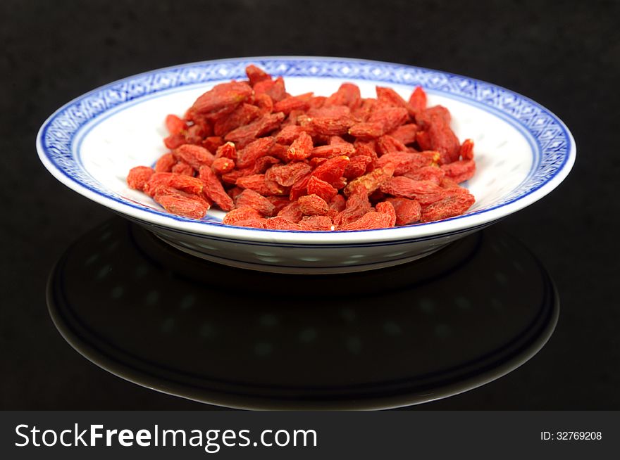 The photograph shows the goji berry placed on a porcelain saucer placed on a black mirrored background. The photograph shows the goji berry placed on a porcelain saucer placed on a black mirrored background.