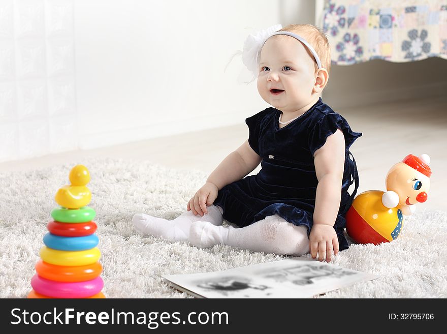 Little cute baby in dress sits on soft carpet among toys and smiles.