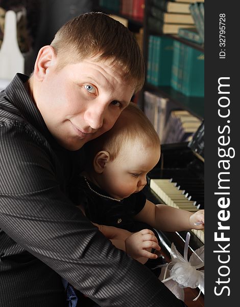 Cute baby sits at knees of her father and touches piano in room. Focus on baby.