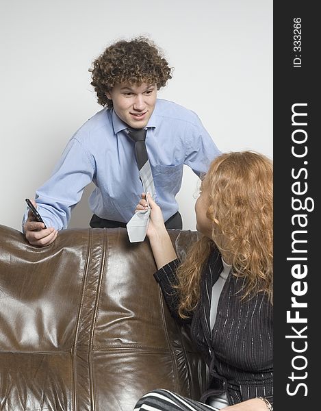 Girl sitting on a leather couch is turning around to a boy standing behind the couch holding a palmtop and is pulling him toward her by his tie. (Boy in relaxed office attire). Girl sitting on a leather couch is turning around to a boy standing behind the couch holding a palmtop and is pulling him toward her by his tie. (Boy in relaxed office attire)