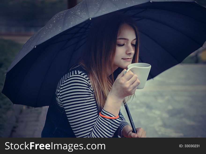 Beautiful girl during a rain under umbrella drinks hot coffee outdoor