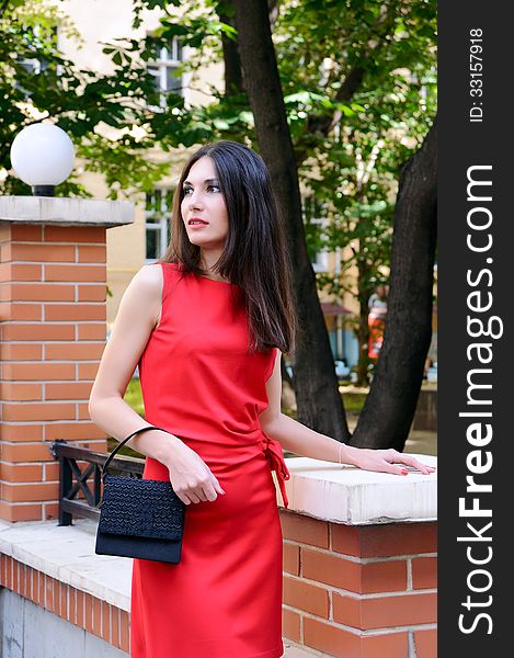 The girl in a red dress stands near a stone fence half-turned