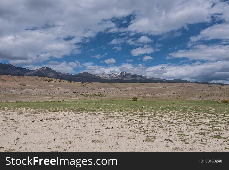 Steppe desert landscape with mountains against the background of blue sky with clouds. Dry land with rare plants as foreground and the mountains, the sky and clouds as a background. Steppe desert landscape with mountains against the background of blue sky with clouds. Dry land with rare plants as foreground and the mountains, the sky and clouds as a background.