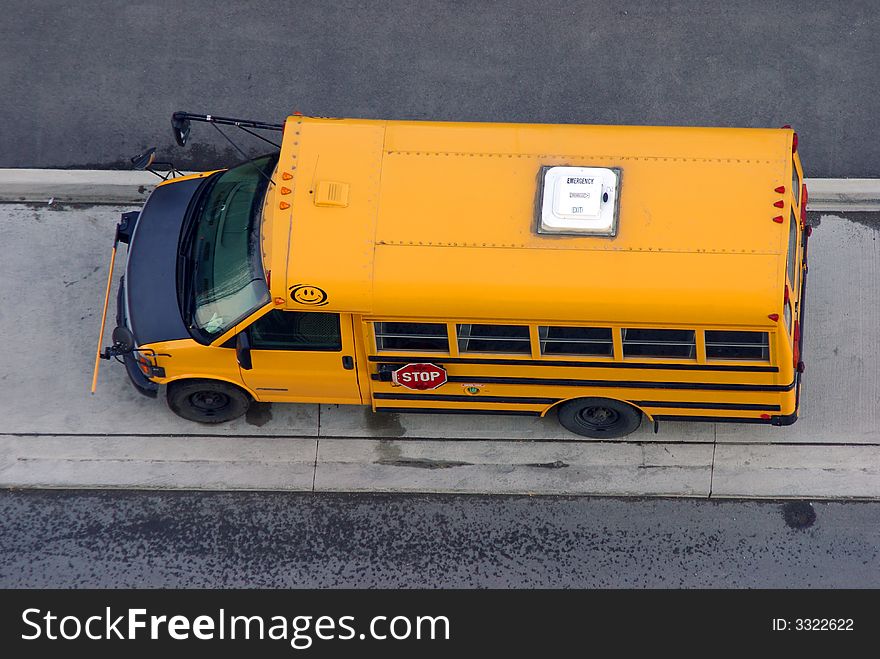 A small parked (van based) yellow school bus from overhead.  Side 'STOP' sign, front safety arm, and room EMERGENCY EXIT visible.