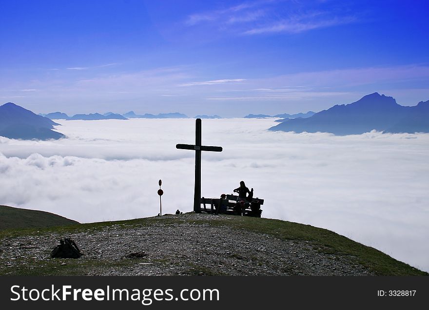 Hikers sitting on a bench close to a summit cross, high above the cloud cover stretching to the background with distant mountain peaks. Hikers sitting on a bench close to a summit cross, high above the cloud cover stretching to the background with distant mountain peaks