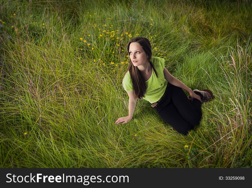 Long hair summer girl wearing green t-shirt and black trousers is sitting in the grass and looking up. Long hair summer girl wearing green t-shirt and black trousers is sitting in the grass and looking up.