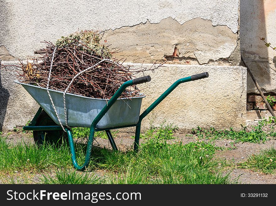 Wheelbarrow filled with cut branches standing in the grass near the old wall. Wheelbarrow filled with cut branches standing in the grass near the old wall