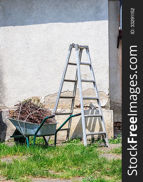 Metal ladder and a wheelbarrow filled with cut branches standing in the grass near the old wall. Metal ladder and a wheelbarrow filled with cut branches standing in the grass near the old wall