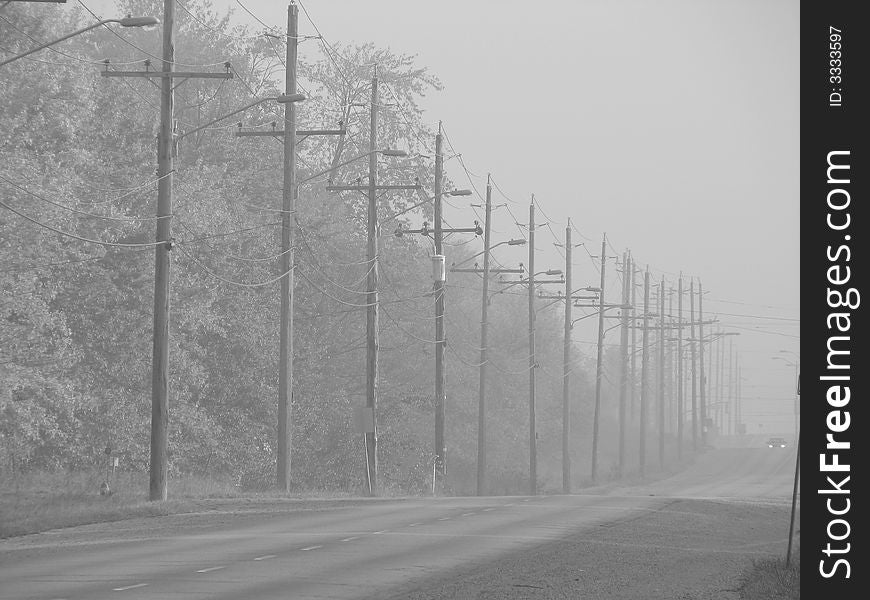 A road in bad foggy weather with a line of electricity poles and the headlights of a car in the horizon. A road in bad foggy weather with a line of electricity poles and the headlights of a car in the horizon