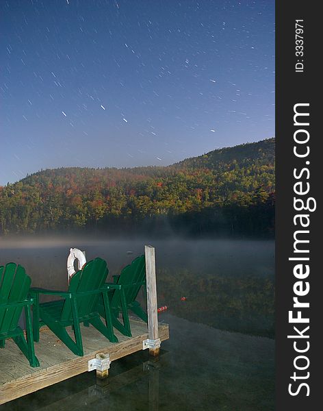 Star trails and fog late at night over a lake in the Adirondack Mountains.  Empty lawn chairs look out at the Autumn foliage. Star trails and fog late at night over a lake in the Adirondack Mountains.  Empty lawn chairs look out at the Autumn foliage.