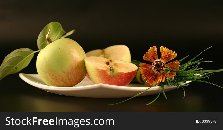 Plate with apples and a bunch of a grass with a flower on a black background.