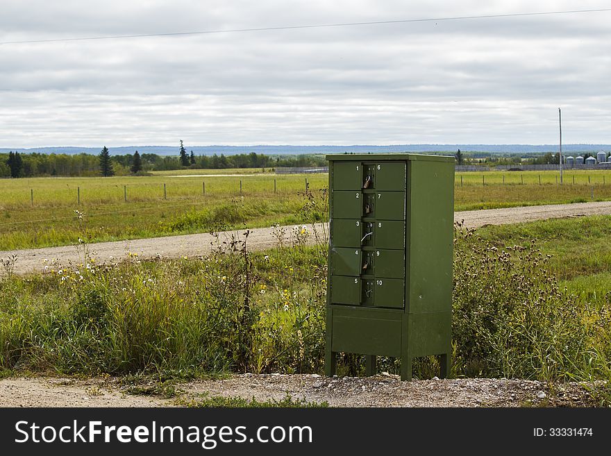 A green metal mailbox with ten boxes beside a road in the countryside with trees and hills in the background. A green metal mailbox with ten boxes beside a road in the countryside with trees and hills in the background