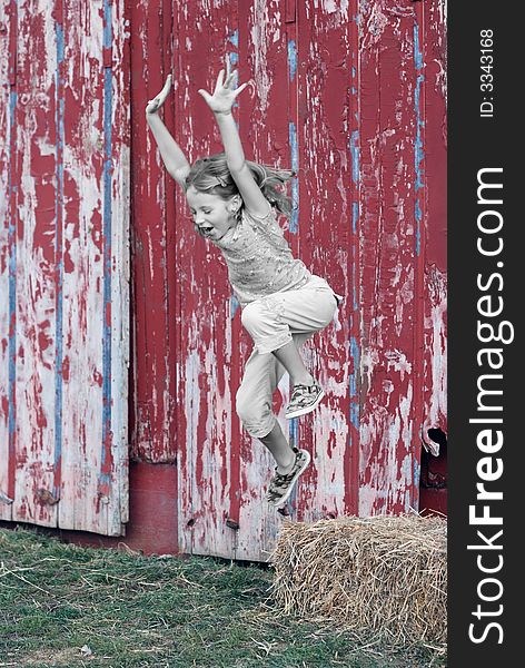 A pretty young girl leaps from a bale of hay with a red barn in the background. A pretty young girl leaps from a bale of hay with a red barn in the background.