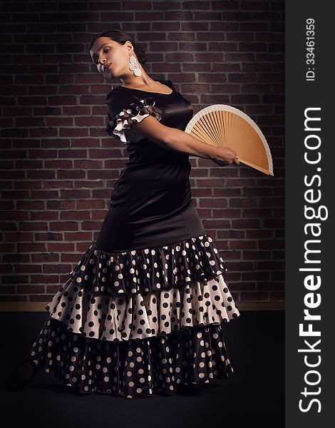 Beautiful young woman wearing brown color silk dress with polka dots is dancing flamenco with spanish hand fan. over red brick wall background. Beautiful young woman wearing brown color silk dress with polka dots is dancing flamenco with spanish hand fan. over red brick wall background.