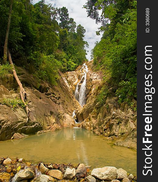 A still shallow pool forming below the cascade of a waterfall in the background deep in a tropical jungle. A still shallow pool forming below the cascade of a waterfall in the background deep in a tropical jungle