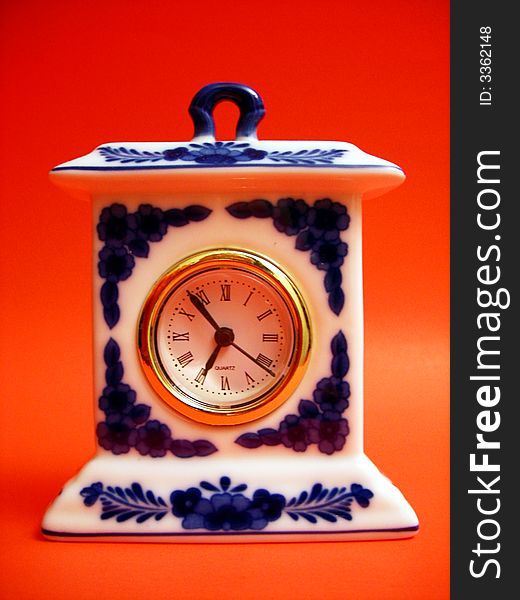 A porcelain clock with blue flowers on a white background. The clock face is in roman numerals and is surrounded by a brass bezel. Background is orange.