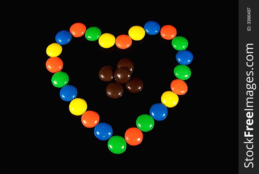 Colorful chocolate candies arranged in a heart shape around a few brown pieces of candy on a black background. Colorful chocolate candies arranged in a heart shape around a few brown pieces of candy on a black background.