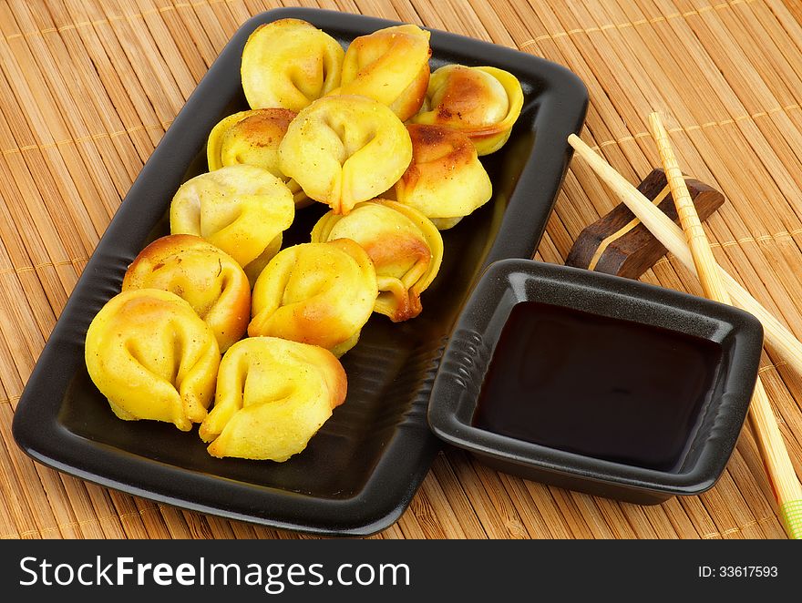 Delicious Fried Crispy Dumplings on Black Plate with Soy Sauce and Chopsticks closeup on Straw mat background