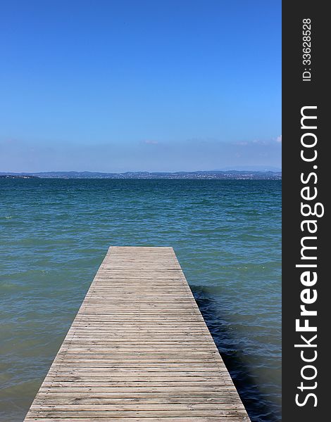 Looking along a wooden jetty out into a lake (Lake Garda in Italy) on a sunny summer day with the other side of the lake visible in the distance.). Looking along a wooden jetty out into a lake (Lake Garda in Italy) on a sunny summer day with the other side of the lake visible in the distance.)