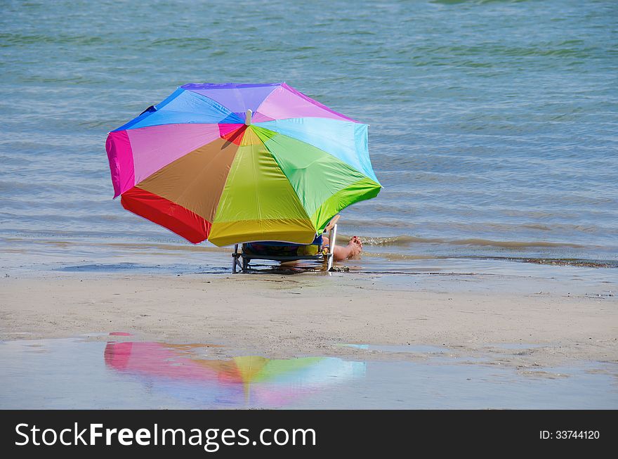 A woman in a beach chair with a very colorful umbrella if sitting down by the water's edge at the beach, the colorful umbrella is reflected in a pool of water. A woman in a beach chair with a very colorful umbrella if sitting down by the water's edge at the beach, the colorful umbrella is reflected in a pool of water.