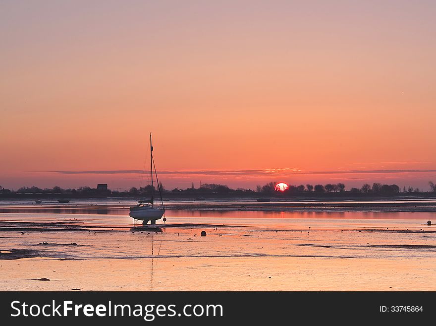 Sunrise over Heybridge Basin Essex UK. Globe of the sun appearing through the trees. Reflection on the water. Solitary yacht in foreground.