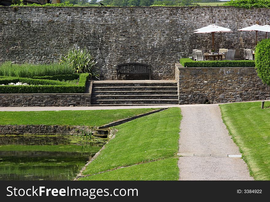 Ancient walled garden in summer, with outdoor patio dining area with tables, chairs and sun umbrellas, a bench, areas of lawn, shrubs and flowers and a pond.