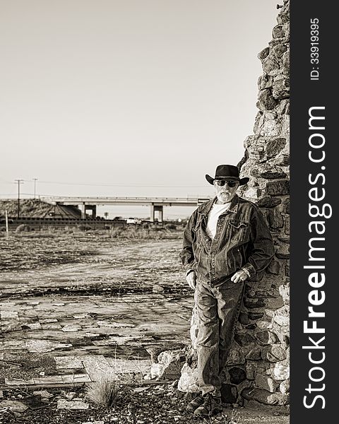 Handsome older man with a gray beard wearing a black cowboy hat leaning against an old stone chimney with a freeway and overpass in the background. Handsome older man with a gray beard wearing a black cowboy hat leaning against an old stone chimney with a freeway and overpass in the background