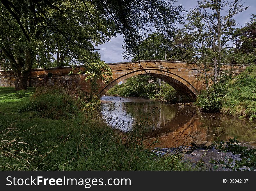 View of the River Esk running under the stone bridge at Lealholm. The River Esk is used for angling. Summer scene Overhanging trees water running over rocks. Sun and shade. View of the River Esk running under the stone bridge at Lealholm. The River Esk is used for angling. Summer scene Overhanging trees water running over rocks. Sun and shade.