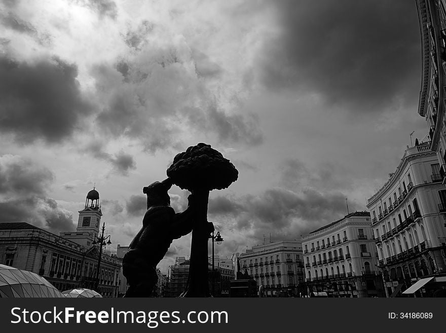 Wide-angle black and white shot of Madrid's Puerta del Sol ('Gate of the Sun') on a cloudy morning. The famous Oso y MadroÃ±o sculpture is to be seen in the foreground, with the Old Post Office Tower and the new glass entrance to the Metro & CercanÃ­as station in the background. Wide-angle black and white shot of Madrid's Puerta del Sol ('Gate of the Sun') on a cloudy morning. The famous Oso y MadroÃ±o sculpture is to be seen in the foreground, with the Old Post Office Tower and the new glass entrance to the Metro & CercanÃ­as station in the background.