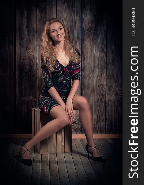 Attractive happy smiling woman with blond long curly hair and fine figure is wearing modern short dress, high-heel shoes and is sitting in a pose on wooden box. Over wooden wall background. Attractive happy smiling woman with blond long curly hair and fine figure is wearing modern short dress, high-heel shoes and is sitting in a pose on wooden box. Over wooden wall background.