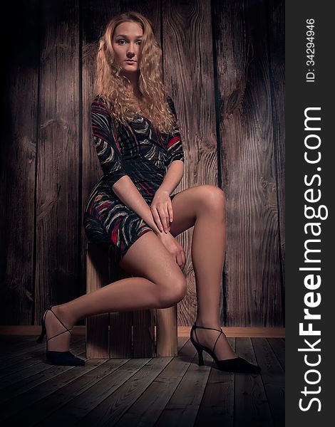 Gorgeous woman with blond long curly hair and fine figure is wearing modern short dress, high-heel shoes and is sitting in a pose on wooden box. Over wooden wall background. Gorgeous woman with blond long curly hair and fine figure is wearing modern short dress, high-heel shoes and is sitting in a pose on wooden box. Over wooden wall background.