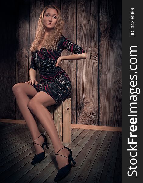 Gorgeous woman with blond long curly hair and fine figure is wearing modern short dress, high-heel shoes and is sitting in a pose on wooden box. Over wooden wall background. Gorgeous woman with blond long curly hair and fine figure is wearing modern short dress, high-heel shoes and is sitting in a pose on wooden box. Over wooden wall background.