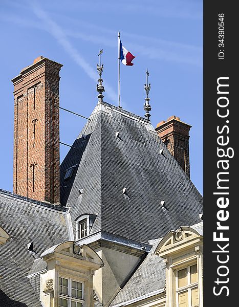 Detail of the roof of the castle of Fontainebleau. The flag of France is silhouetted against a blue sky. Detail of the roof of the castle of Fontainebleau. The flag of France is silhouetted against a blue sky.