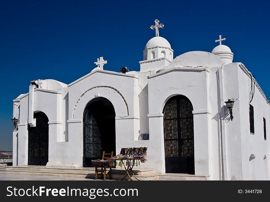 A white Greek orthodox church on a clear blue sky background with a trinket stall.