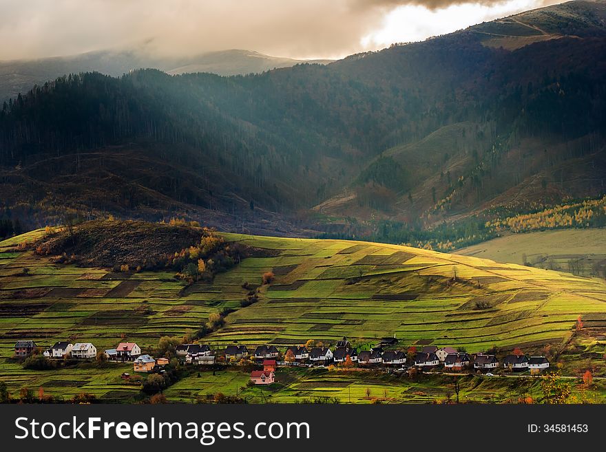 Autumn landscape. village on the hillside. forest on the mountain covered with red and yellow leaves. over the mountains the beam of light falls on a clearing at the top of the hill. Autumn landscape. village on the hillside. forest on the mountain covered with red and yellow leaves. over the mountains the beam of light falls on a clearing at the top of the hill.