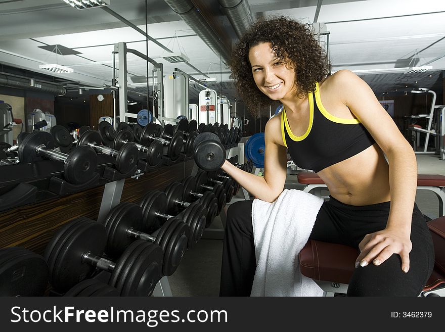 Young woman working out in gym with dumbbell. Smiling and looking at camera. Front view. Young woman working out in gym with dumbbell. Smiling and looking at camera. Front view