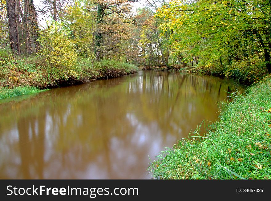 The photograph shows a small river flowing through the forest. It is autumn, the leaves are green, yellow and brown. The photograph shows a small river flowing through the forest. It is autumn, the leaves are green, yellow and brown.