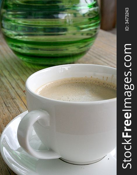 Close-up of a white coffee cup on a timber table with a green glass vase in the background. Close-up of a white coffee cup on a timber table with a green glass vase in the background