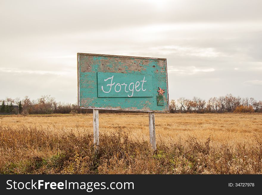 A green wood sign with peeling paint announcing the name of a ghost town as "Forget" with a small lily in the corner. A green wood sign with peeling paint announcing the name of a ghost town as "Forget" with a small lily in the corner