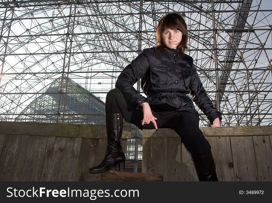 Tough young girl in black clothes on old wooden fence. Construction site at background. Tough young girl in black clothes on old wooden fence. Construction site at background