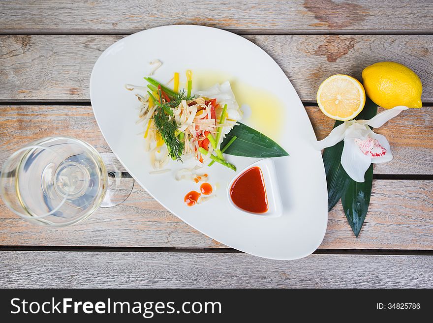 Serving of steamed sea bass fillet on a plate in a restaurant, on a wooden table with white wine and decor. Serving of steamed sea bass fillet on a plate in a restaurant, on a wooden table with white wine and decor