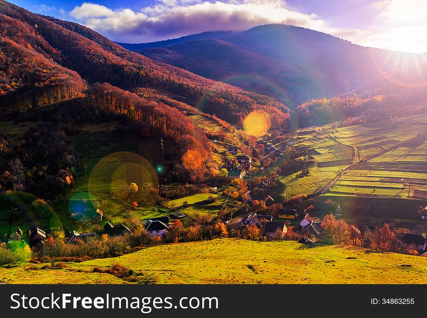 Autumn landscape. village on the hillside. forest on the mountain covered with red and yellow leaves. over the mountains the beam of light falls on a clearing at the top of the hill. Autumn landscape. village on the hillside. forest on the mountain covered with red and yellow leaves. over the mountains the beam of light falls on a clearing at the top of the hill.